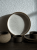 A large ceramic plate with a wavy, textured sand pattern leans against a white textured wall. It is surrounded by a matching dark brown tableware set, including small cups, bowls, and saucers under dramatic sunlight.