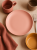 A top-down view of a matte, pastel pink ceramic plate centered on a beige surface. The warm-toned table setting includes a terracotta-colored mug, a mustard yellow bowl, a beige cup, a linen napkin, and a silver fork.