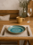 A turquoise ceramic plate with a swirl pattern sits on a linen placemat flanked by a fork and knife with wooden handles. The rustic table setting includes a yellow mug,  wine glass, and a small glass vase in the background.