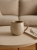 A speckled beige ceramic mug sits on a round wooden coffee table next to a folded napkin and fork. The minimalist setting features a softly blurred, warm cream-colored sofa in the background.