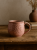 A pink, textured ceramic mug sits on a rustic wooden table. In the softly blurred background, a stack of books and a small terracotta vase with dried grass add to the warm, cozy atmosphere.