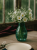 A translucent teal glass vase filled with white wildflowers and baby's breath sits on a rustic wooden table. The vase is part of a cozy table setting next to a window, accompanied by a plate and a fork.
