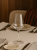 A clear wine glass with a thin stem sits empty on a set table. The setting includes plates and cutlery on a beige tablecloth.
